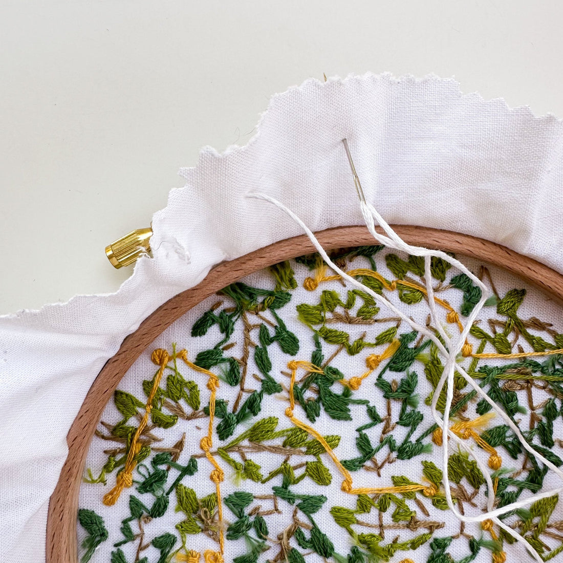 Embroidery hoop with green and yellow leaf design, white fabric, and a needle with white thread.