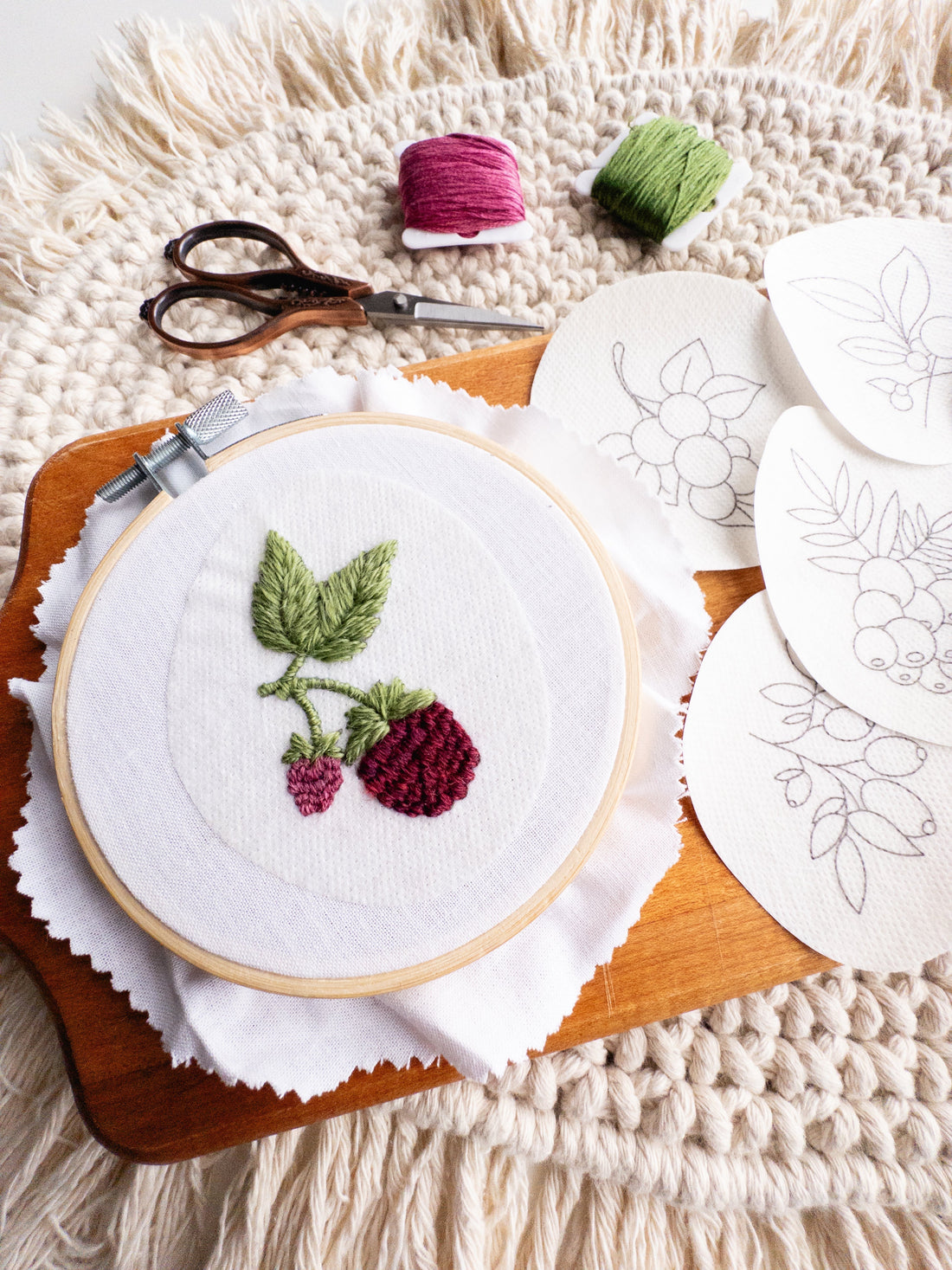 Embroidery hoop with raspberry design, thread, scissors, and stick and stitch patterns on a wooden board.