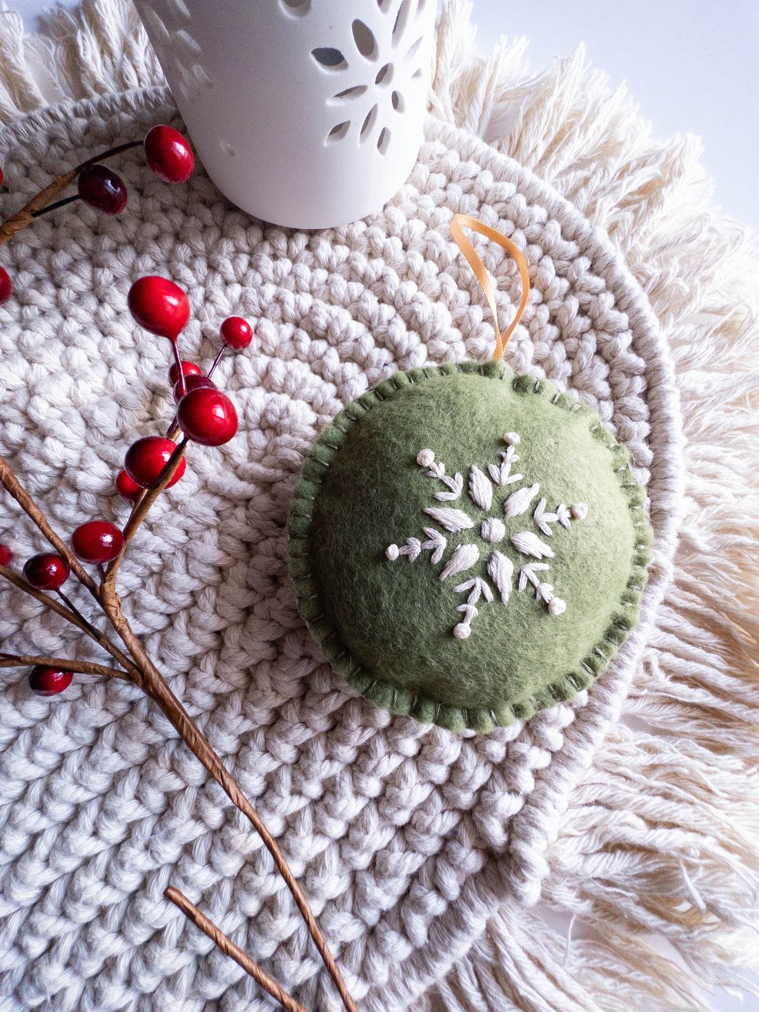 Green felt ornament with embroidered white snowflake, red berries, and white candle holder on a macrame mat.
