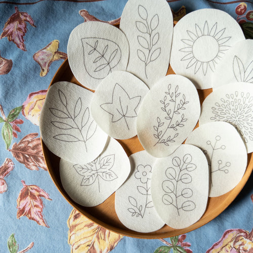 Autumn botanical embroidery transfer patterns on water-soluble paper, featuring leaves, flowers, and a sun, on a wooden tray.