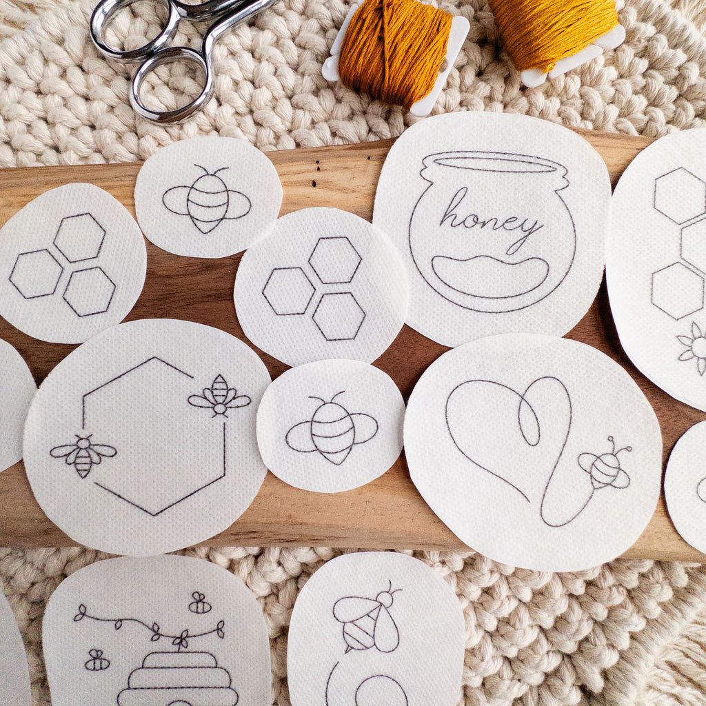 White water-soluble stick and stitch bee embroidery patterns on a wooden board with yarn and scissors.