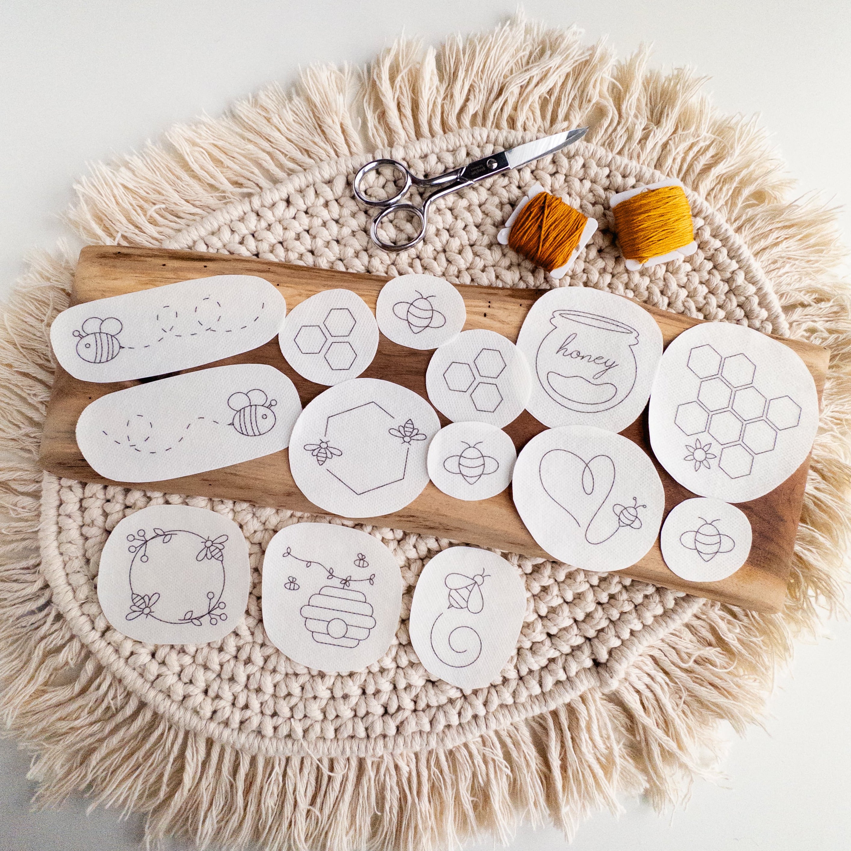 14 white water-soluble stick and stitch bee embroidery patterns on a wooden board with thread and scissors.