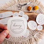 Hand holding a water-soluble stick and stitch honeybee embroidery pattern, surrounded by other bee designs, scissors, and yellow floss.