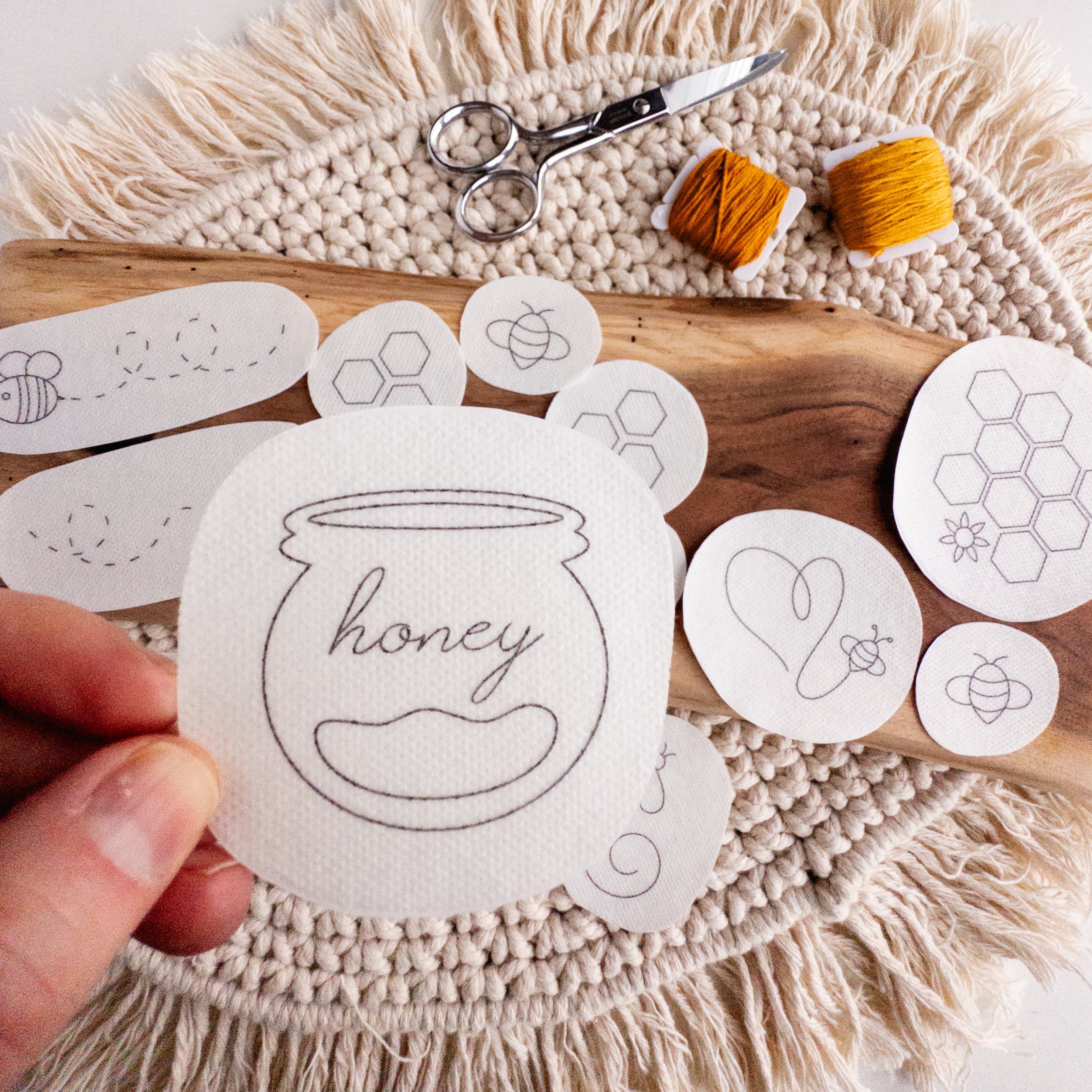Hand holding a water-soluble stick and stitch honeybee embroidery pattern, surrounded by other bee designs, scissors, and yellow floss.