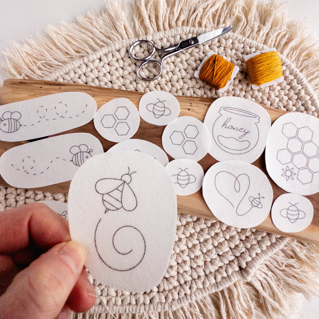Hand holding a white oval stick and stitch bee embroidery pattern, surrounded by other bee and honeycomb designs.