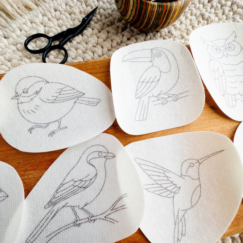 Nine white dissolvable stick and stitch bird embroidery patterns, including an owl, toucan, and hummingbird, on a wooden surface.