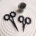 Black floral scissors on a textured white surface with a small container of pins.