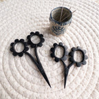 Black floral scissors on a textured white surface with a small container of pins.