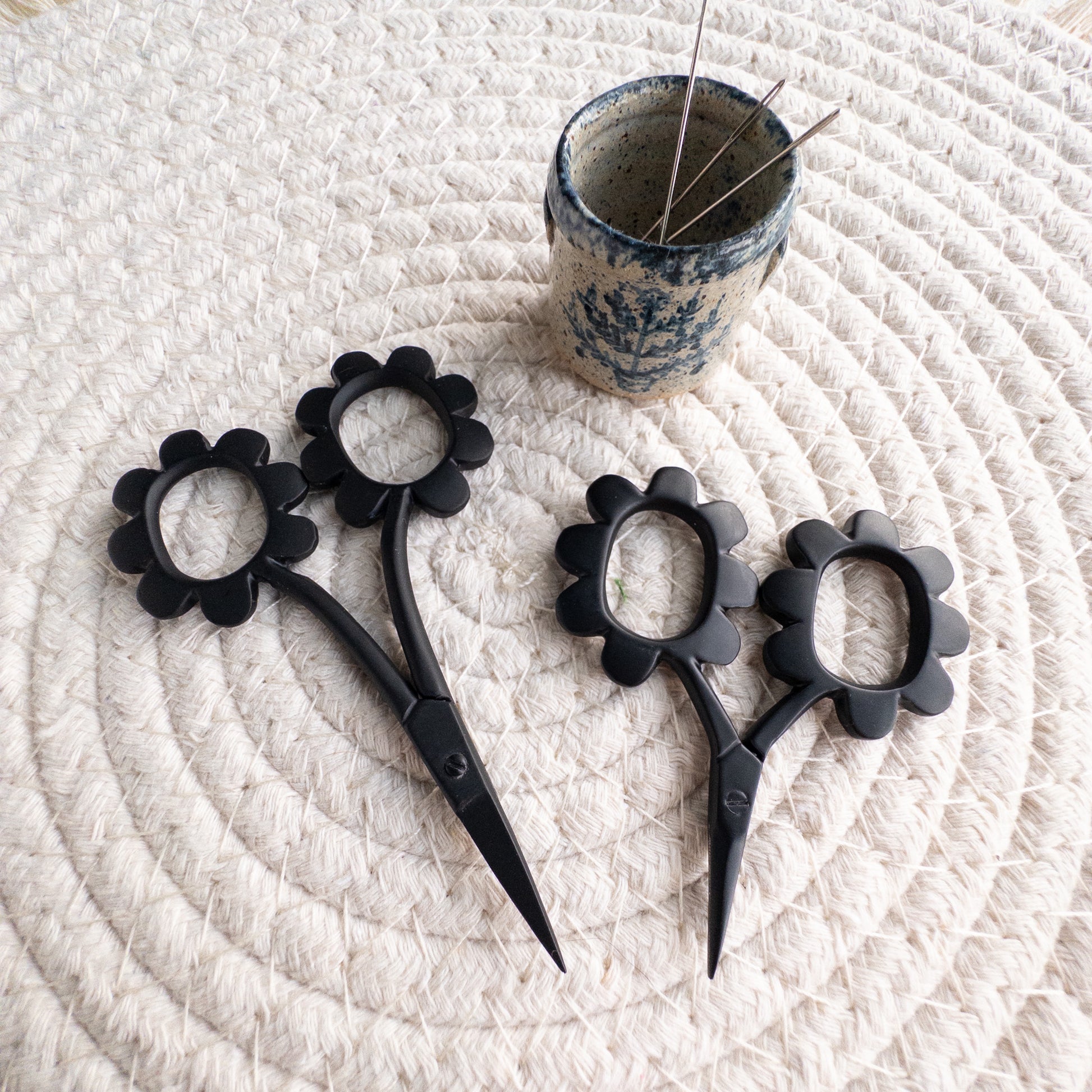 Black floral scissors on a textured white surface with a small container of pins.