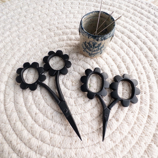 Black floral scissors on a textured white surface with a small container of pins.
