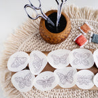 Twelve white water-soluble butterfly embroidery patterns on a wood board, with scissors and floss.