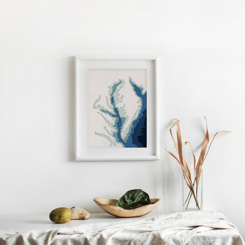 Framed Chesapeake Bay cross-stitch pattern, a minimalist blue bathymetry map on white fabric.