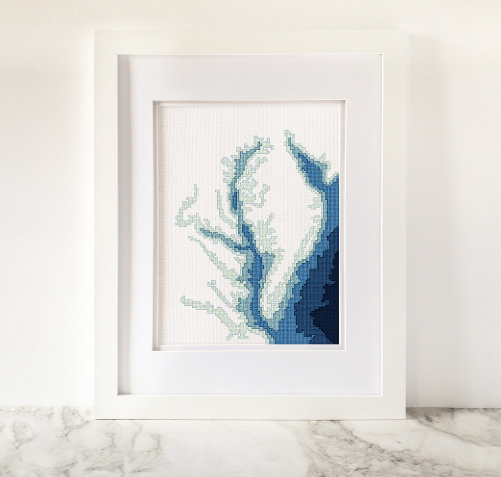 Framed Chesapeake Bay cross-stitch art with blue and green bathymetry map design on white fabric.