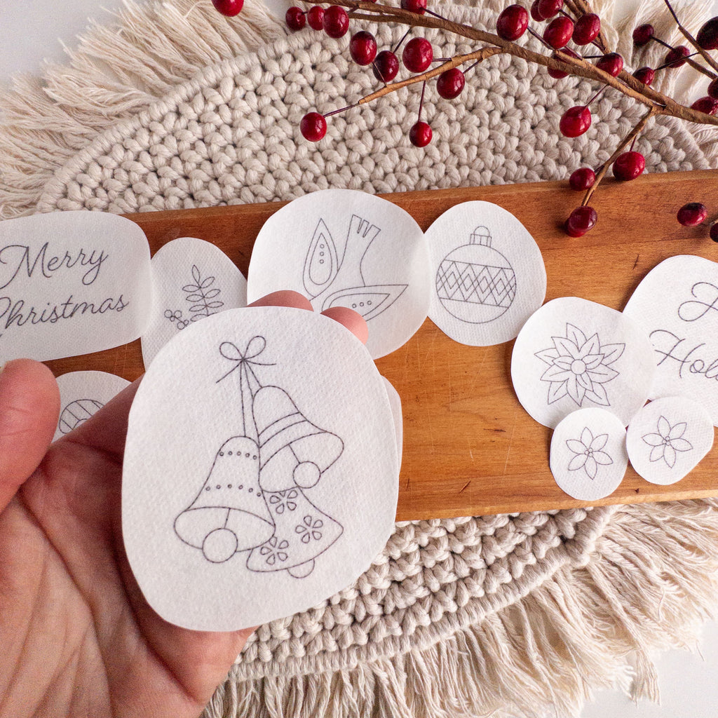 Hand holding a Christmas bells stick and stitch pattern, surrounded by other holiday embroidery designs on a wooden board.