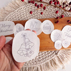 Hand holding a Christmas bells stick and stitch pattern, surrounded by other holiday embroidery designs on a wooden board.