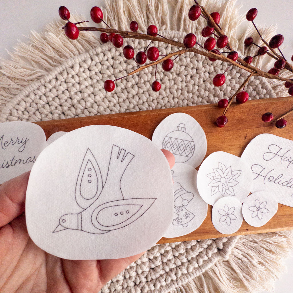 Hand holding a white water-soluble stick and stitch dove pattern, with other Christmas designs on a wooden board.