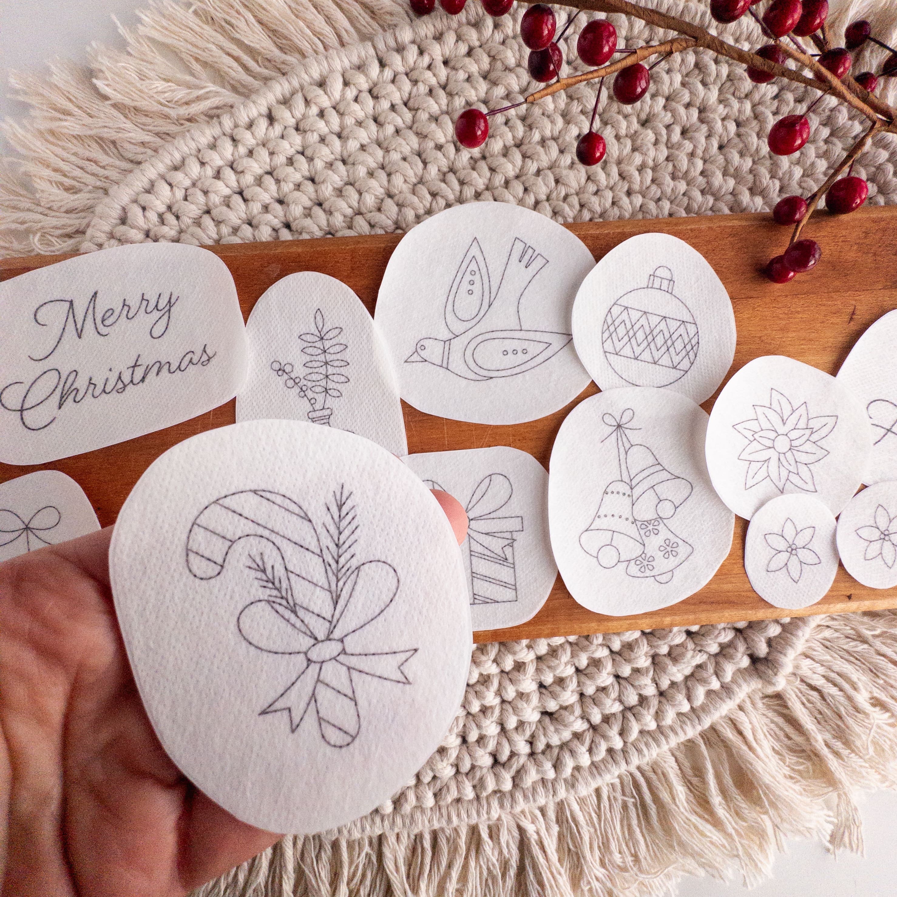 Hand holding a Christmas stick and stitch embroidery pattern of a candy cane and holly, with other holiday designs on a wooden board.