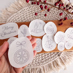 Hand holding a white, oval Christmas ornament stick and stitch pattern, surrounded by 11 other festive designs on a wooden board with red berries.