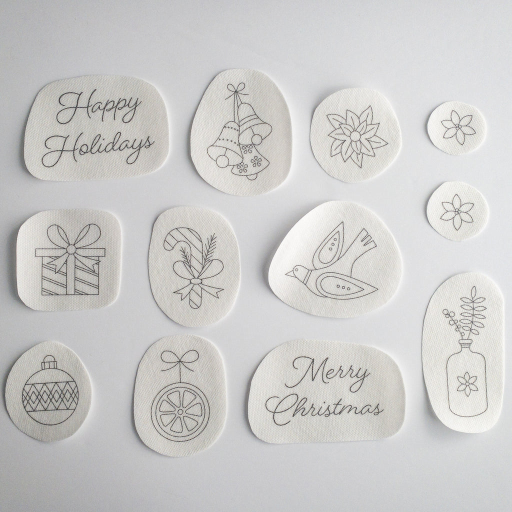 12 white water-soluble stick and stitch Christmas embroidery patterns, including "Happy Holidays," bells, poinsettias, a gift, candy cane, dove, ornaments, "Merry Christmas," and a vase.