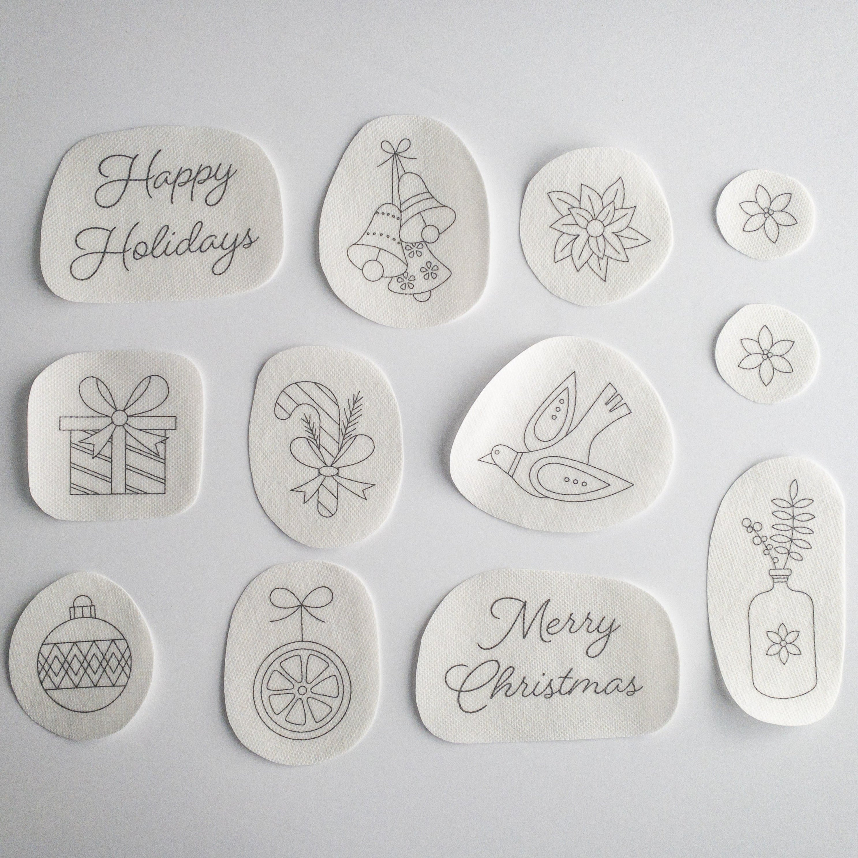 12 white water-soluble stick and stitch Christmas embroidery patterns, including "Happy Holidays," bells, poinsettias, a gift, candy cane, dove, ornaments, "Merry Christmas," and a vase.