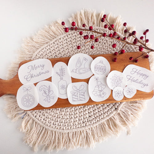 12 white water-soluble Christmas stick and stitch embroidery patterns on a wooden board with red berries.