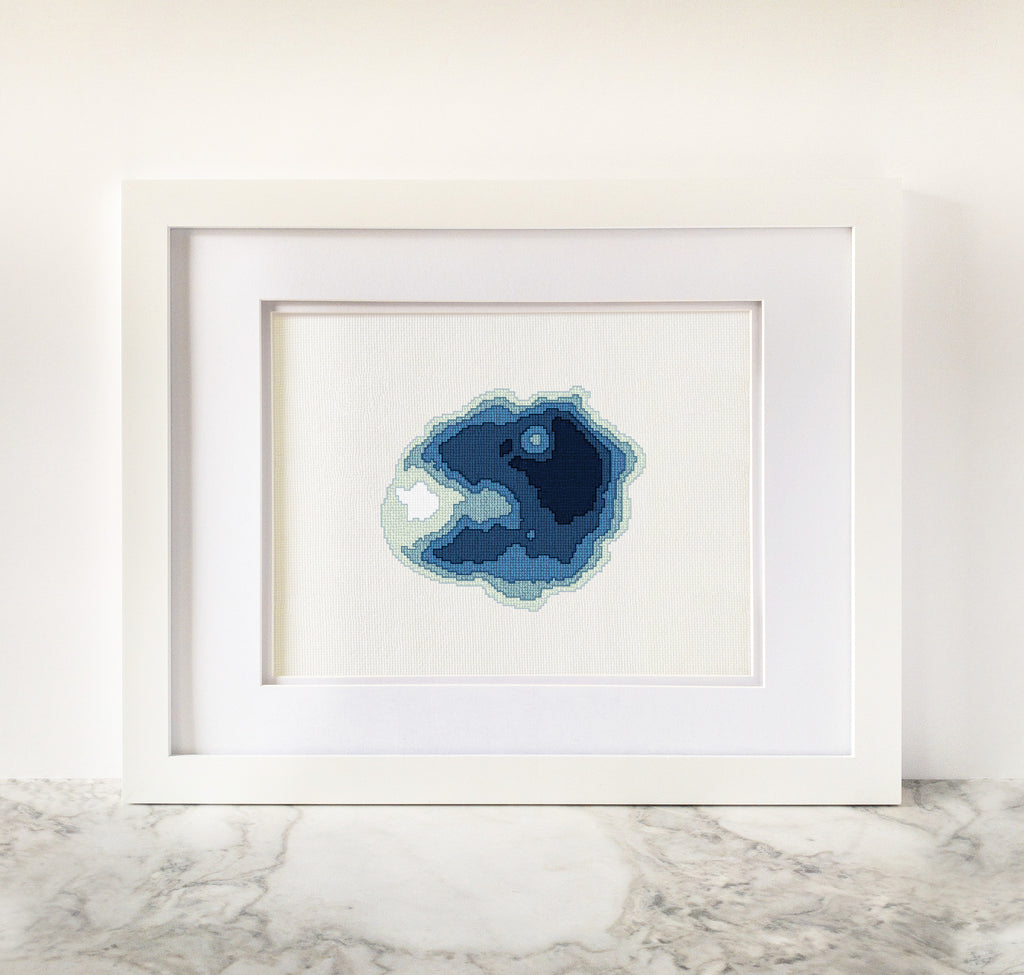 Framed Crater Lake cross-stitch art with a minimalist blue bathymetric map design on white fabric.