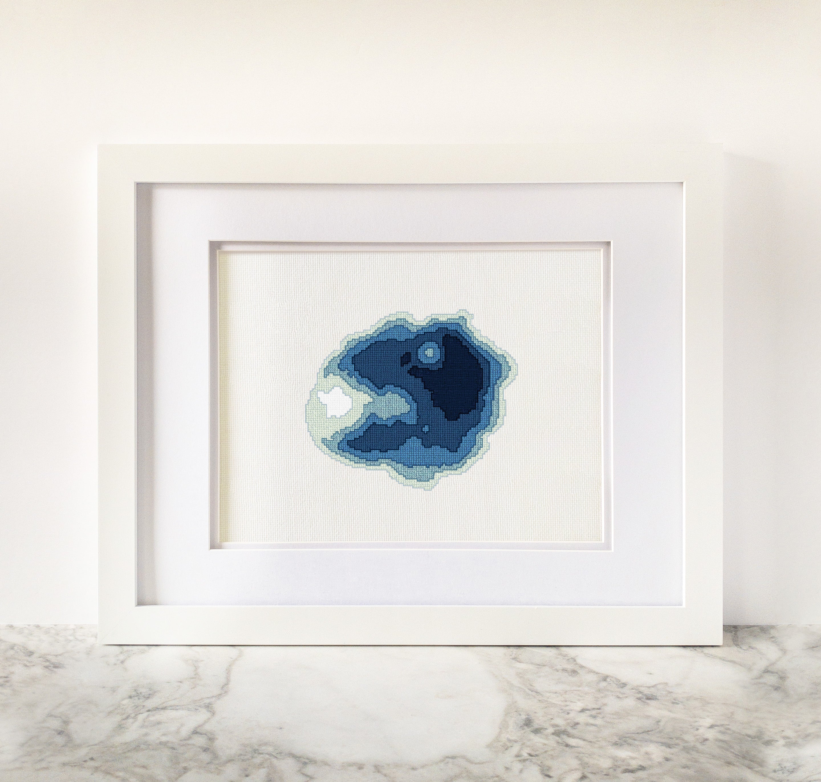 Framed Crater Lake cross-stitch art with a minimalist blue bathymetric map design on white fabric.
