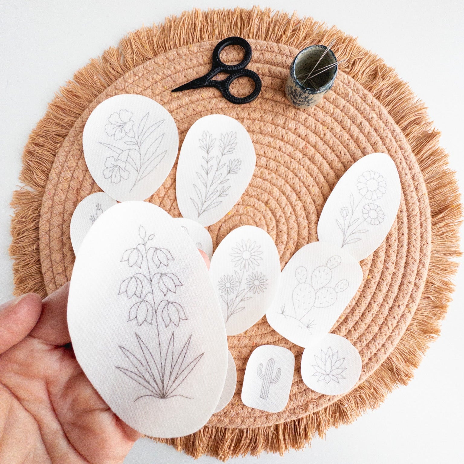 Hand holding a desert botanical embroidery stick and stitch pattern, surrounded by other patterns, scissors, and a needle holder on a woven mat.