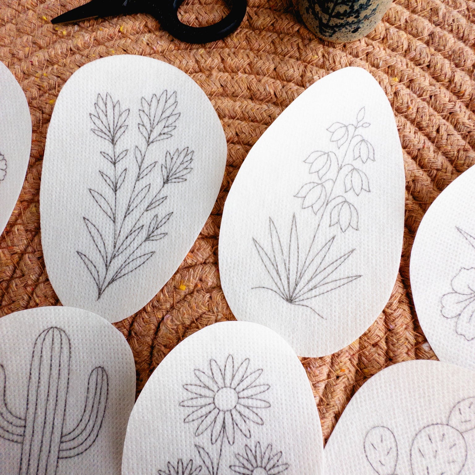 White stick and stitch embroidery patterns featuring desert botanicals like cacti, succulents, and blooms on a woven mat.