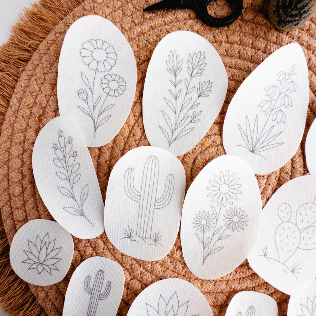 White stick and stitch embroidery patterns featuring 13 desert botanicals like cacti, succulents, and blooms on a woven mat.