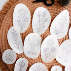 White stick and stitch embroidery patterns featuring 13 desert botanicals like cacti, succulents, and blooms on a woven mat.