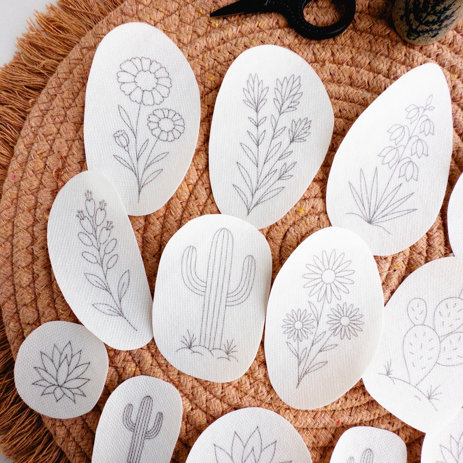 White stick and stitch embroidery patterns featuring 13 desert botanicals like cacti, succulents, and blooms on a woven mat.