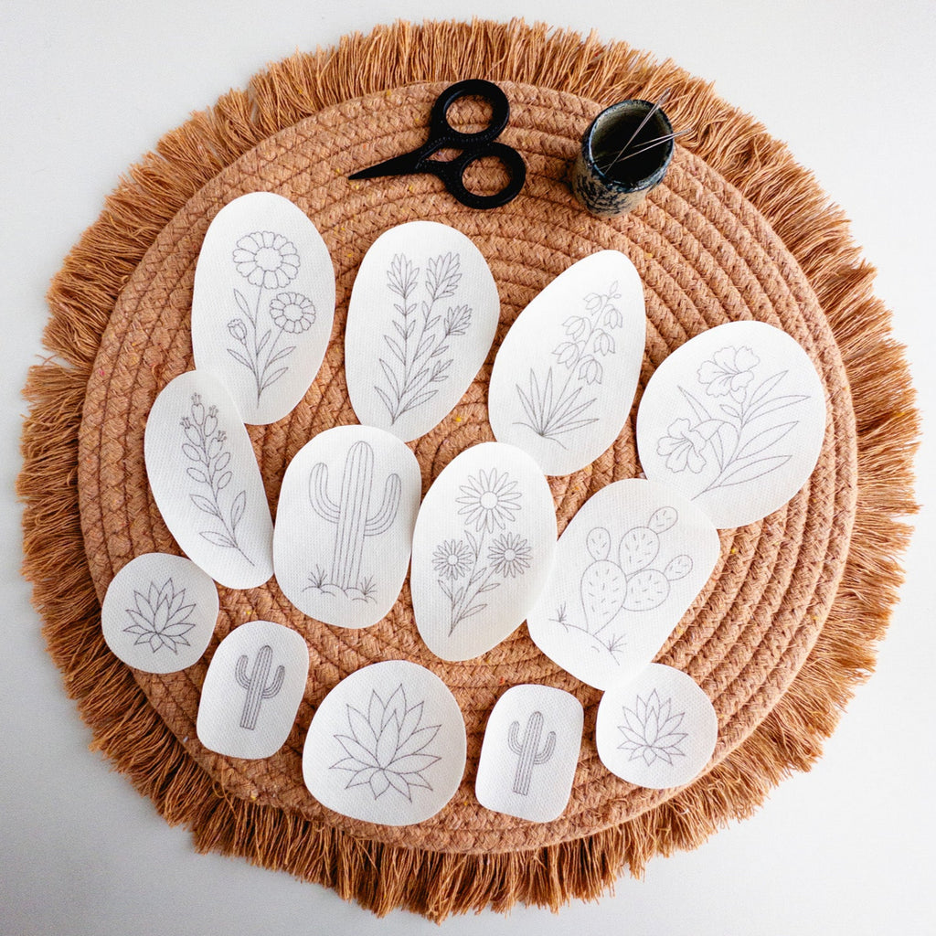 White desert botanical embroidery stick and stitch patterns, including cacti and flowers, on a fringed brown placemat with scissors and needles.