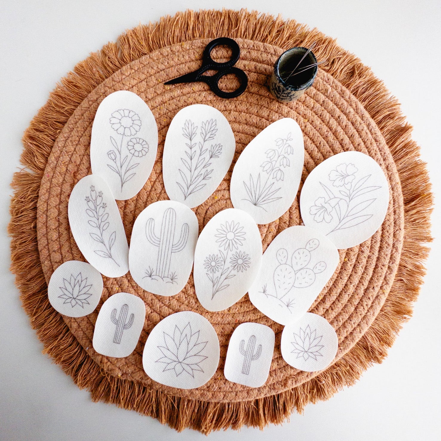 White desert botanical embroidery stick and stitch patterns, including cacti and flowers, on a fringed brown placemat with scissors and needles.