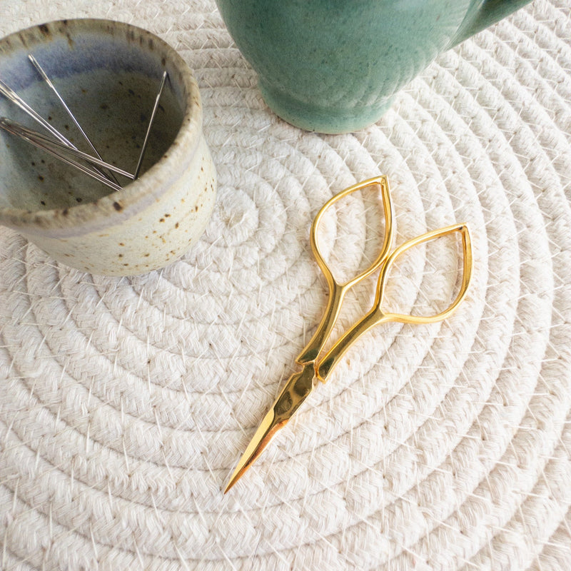 Gold scissors on a textured white surface with a ceramic container and green mug in the background.