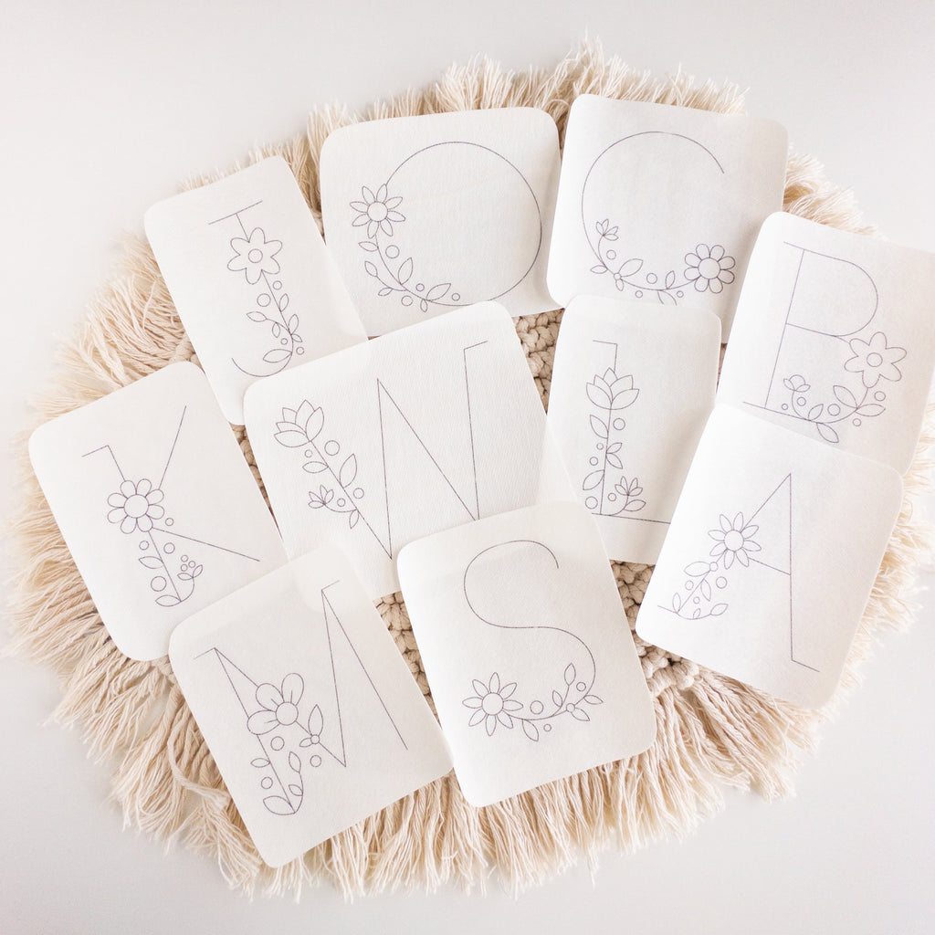 White stick and stitch embroidery patterns featuring large floral letters K, M, N, S, O, C, L, B, A on a macrame mat.