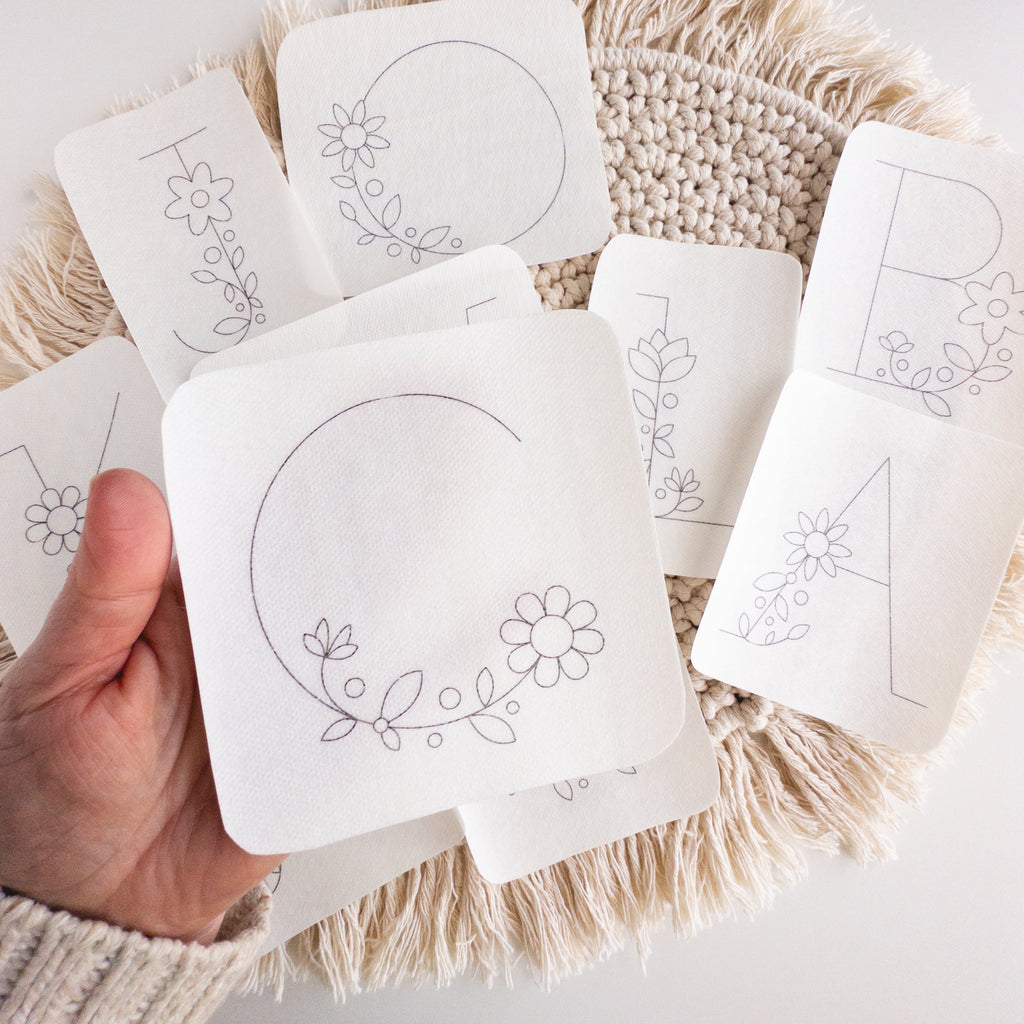 Hand holding white stick and stitch embroidery patterns with floral letters A, B, C, and J designs.