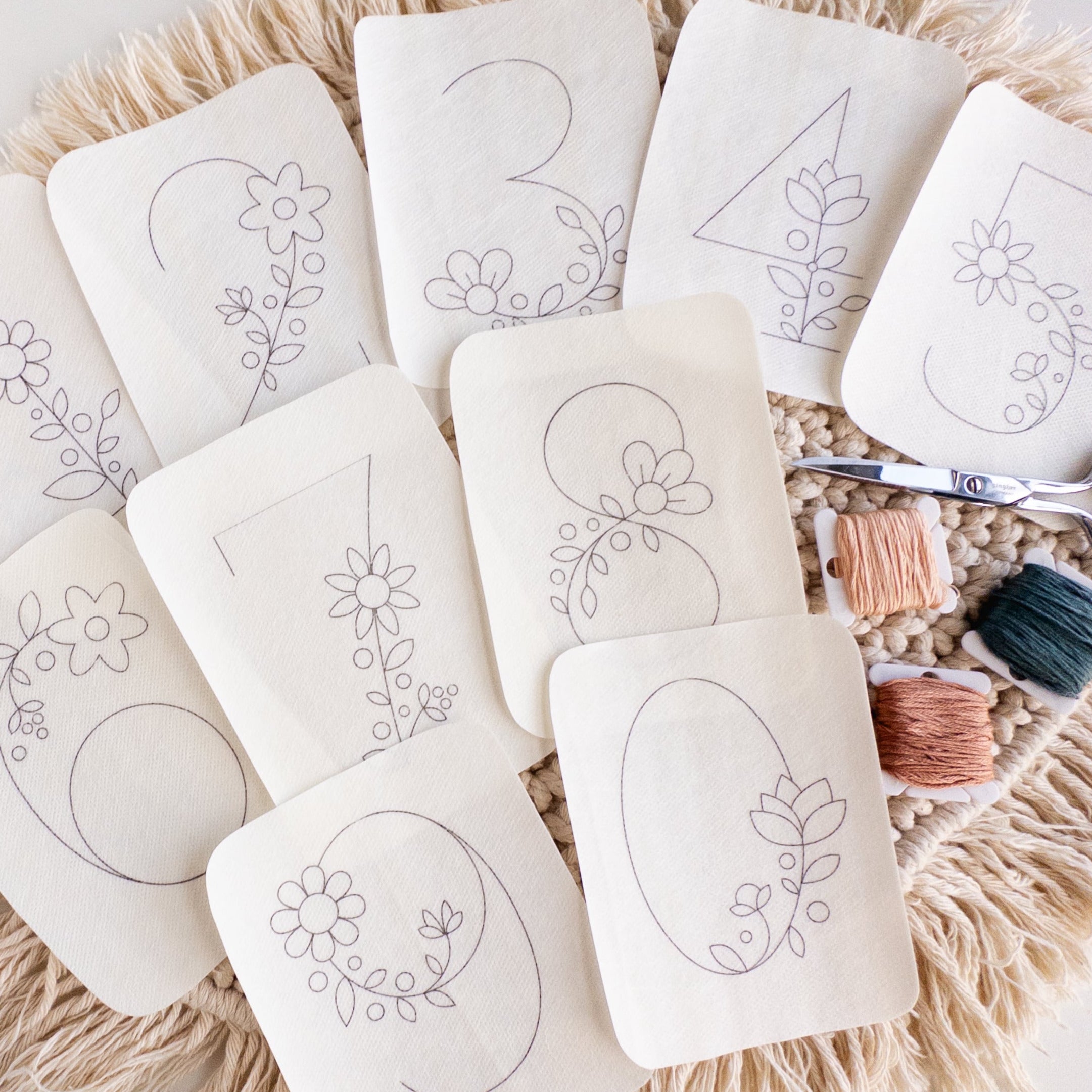 Cream fabric stick and stitch patterns with floral numbers 0-9, embroidery floss, and scissors on a macrame mat.