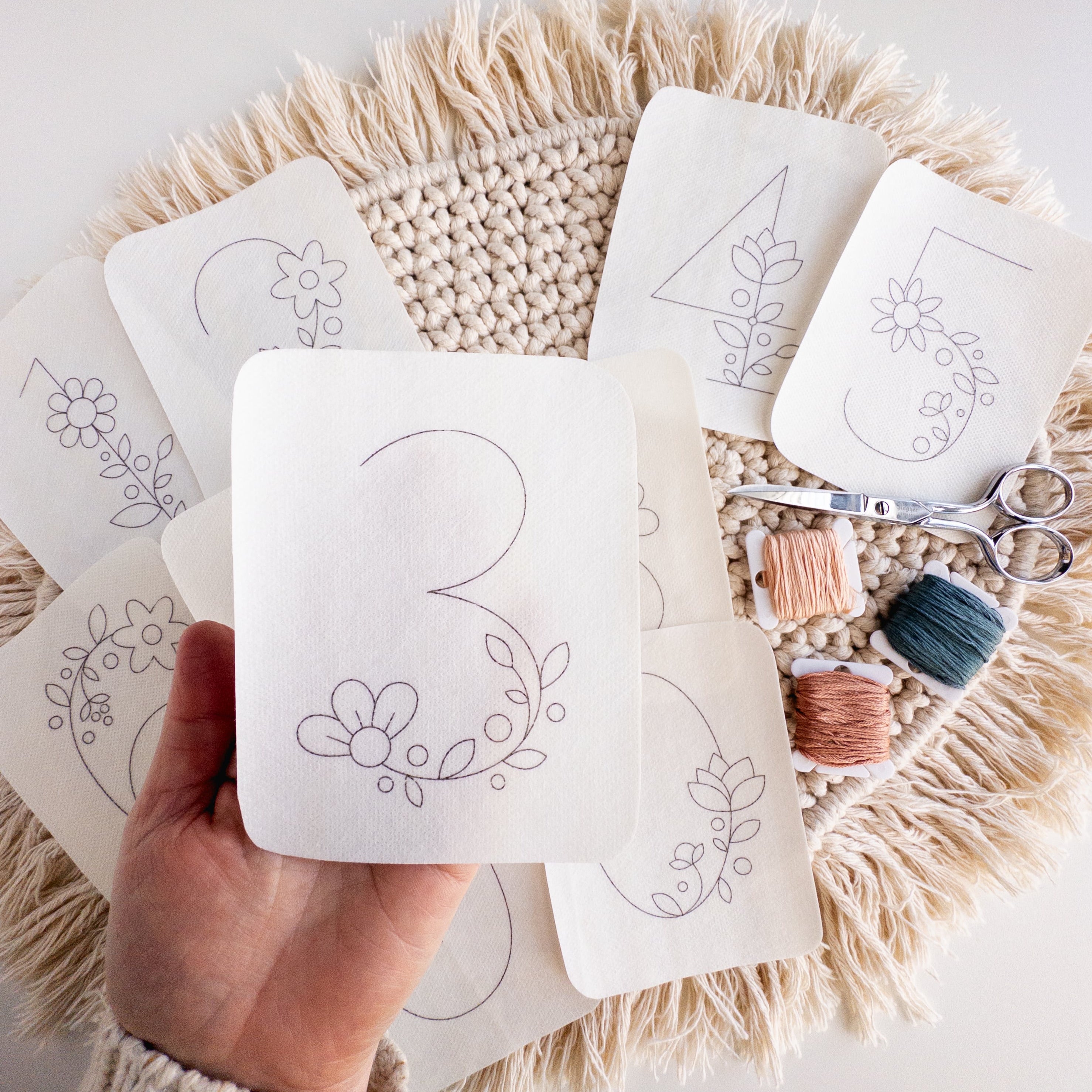 Hand holding a floral number 3 stick and stitch pattern, surrounded by other floral number patterns, embroidery floss, and scissors on a macrame mat.