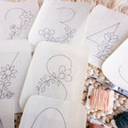 White stick and stitch embroidery patterns with floral numbers, peach floss, and scissors on a textured background.