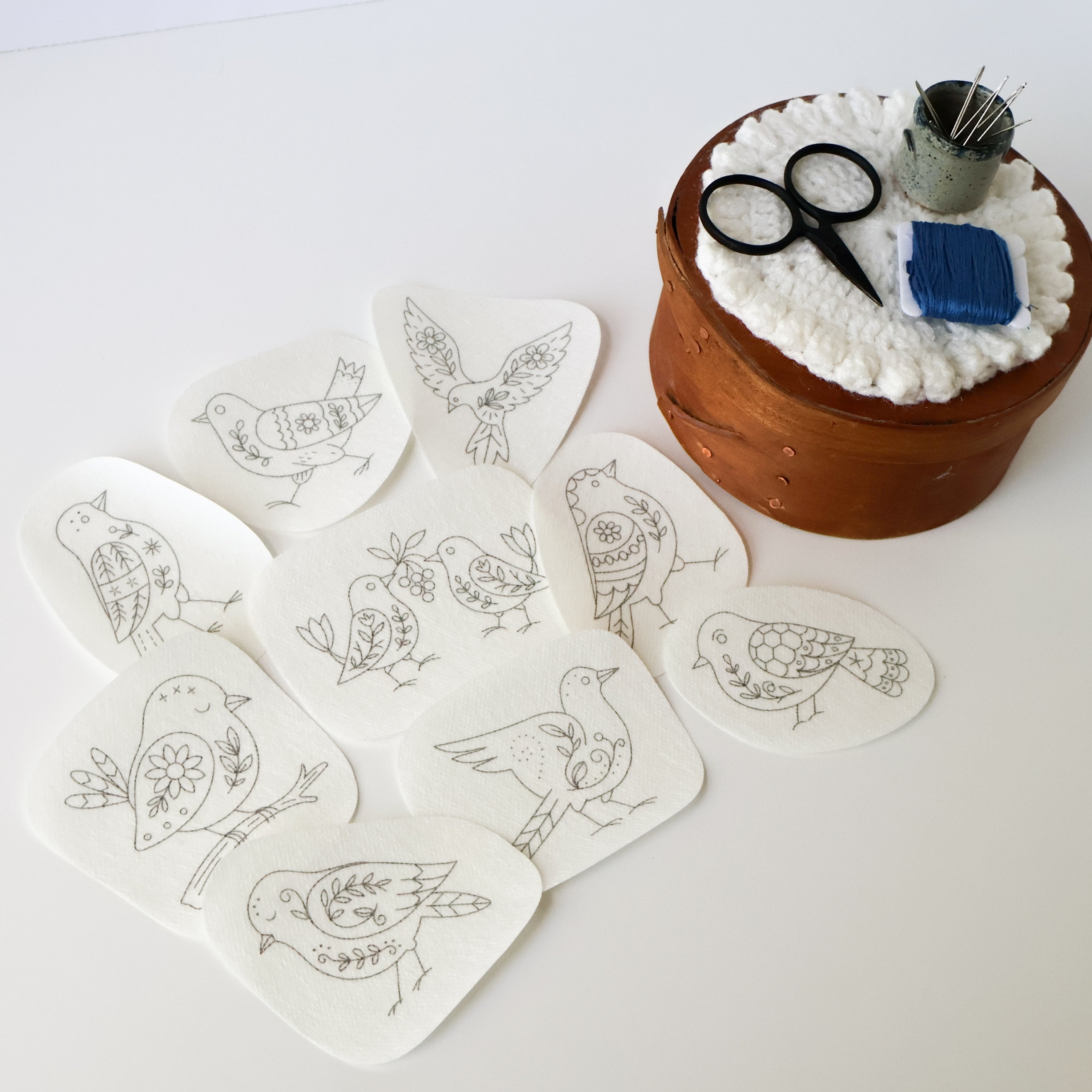 Nine folk art bird embroidery patterns on washaway stabilizer, with scissors, needles, and blue floss.