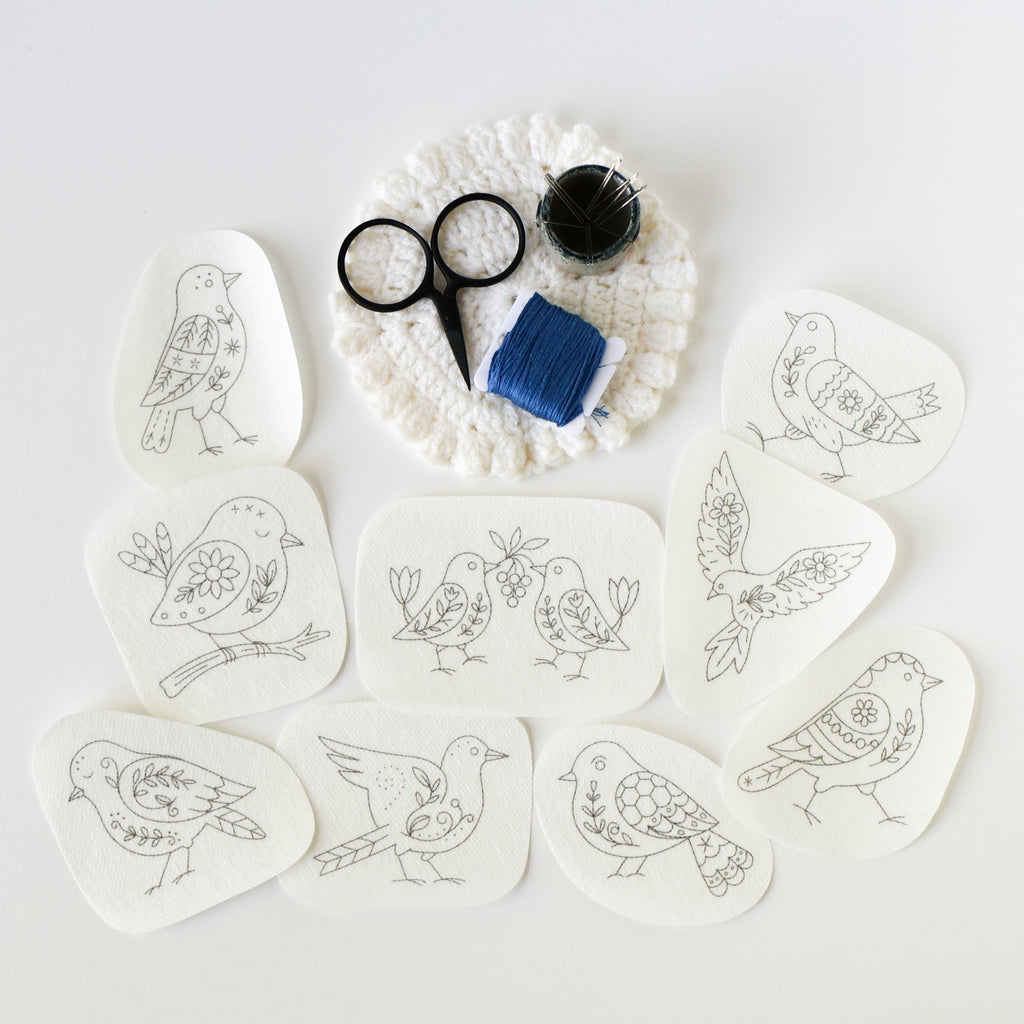 Nine folk bird embroidery patterns on washaway stabilizer, with scissors, blue thread, and pins on a white crochet coaster.