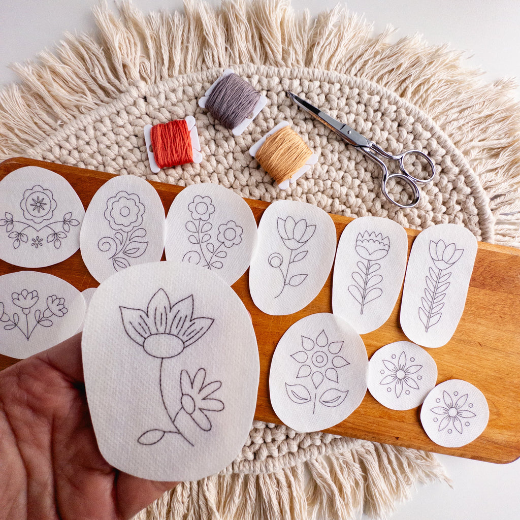 Hand holding a white folk flower embroidery sticker with other floral patterns, thread, and scissors on a wooden board.
