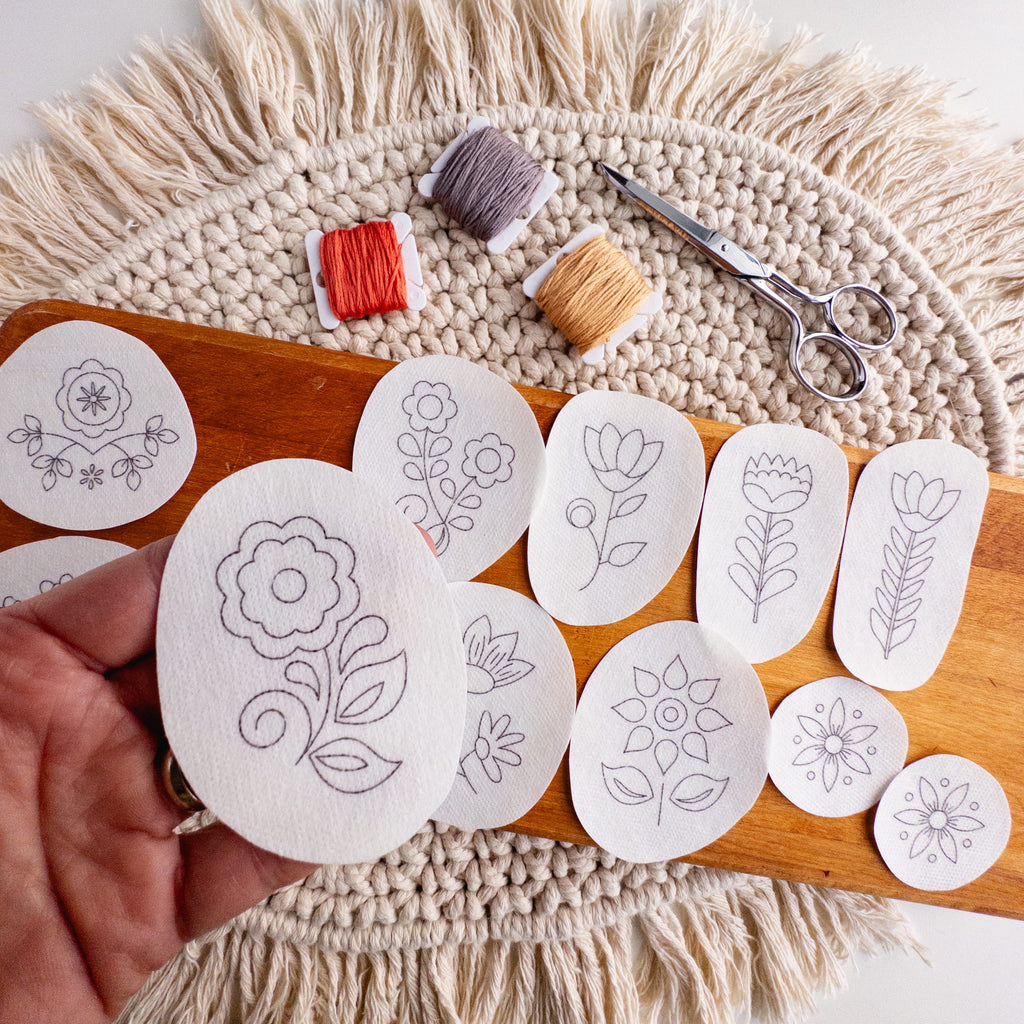 Hand holding a folk flower embroidery pattern sticker, with 11 other Scandinavian designs, thread, and scissors.
