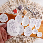Hand holding a folk flower embroidery pattern sticker, with 11 other Scandinavian designs, thread, and scissors.