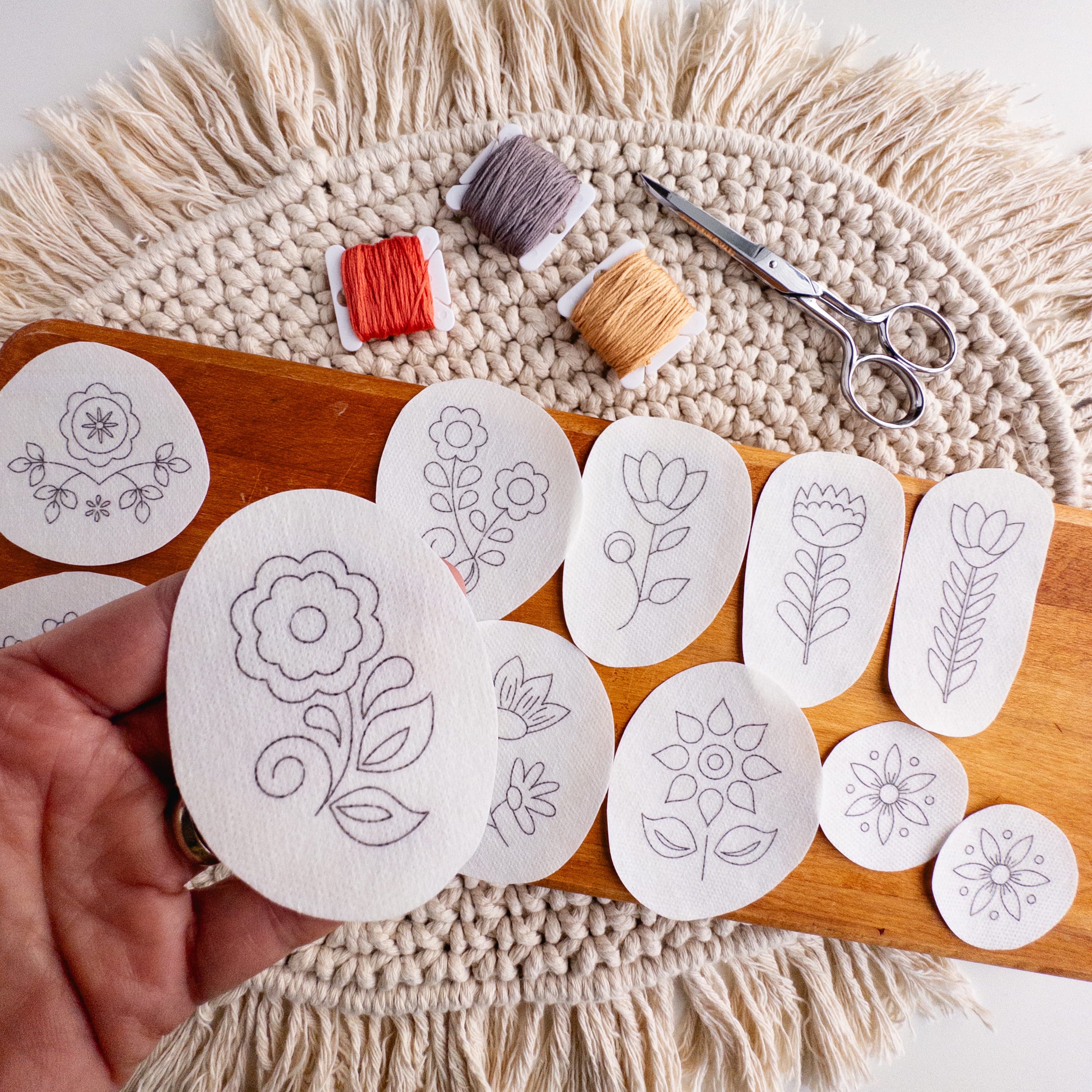 Hand holding a folk flower embroidery pattern sticker, with 11 other Scandinavian designs, thread, and scissors.