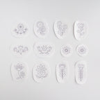 12 white water-soluble stabilizer stickers with purple Scandinavian folk flower embroidery patterns.