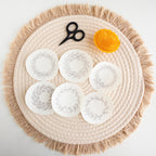 Six round, white wash-away embroidery stickers with delicate fruit wreath designs on a woven placemat.