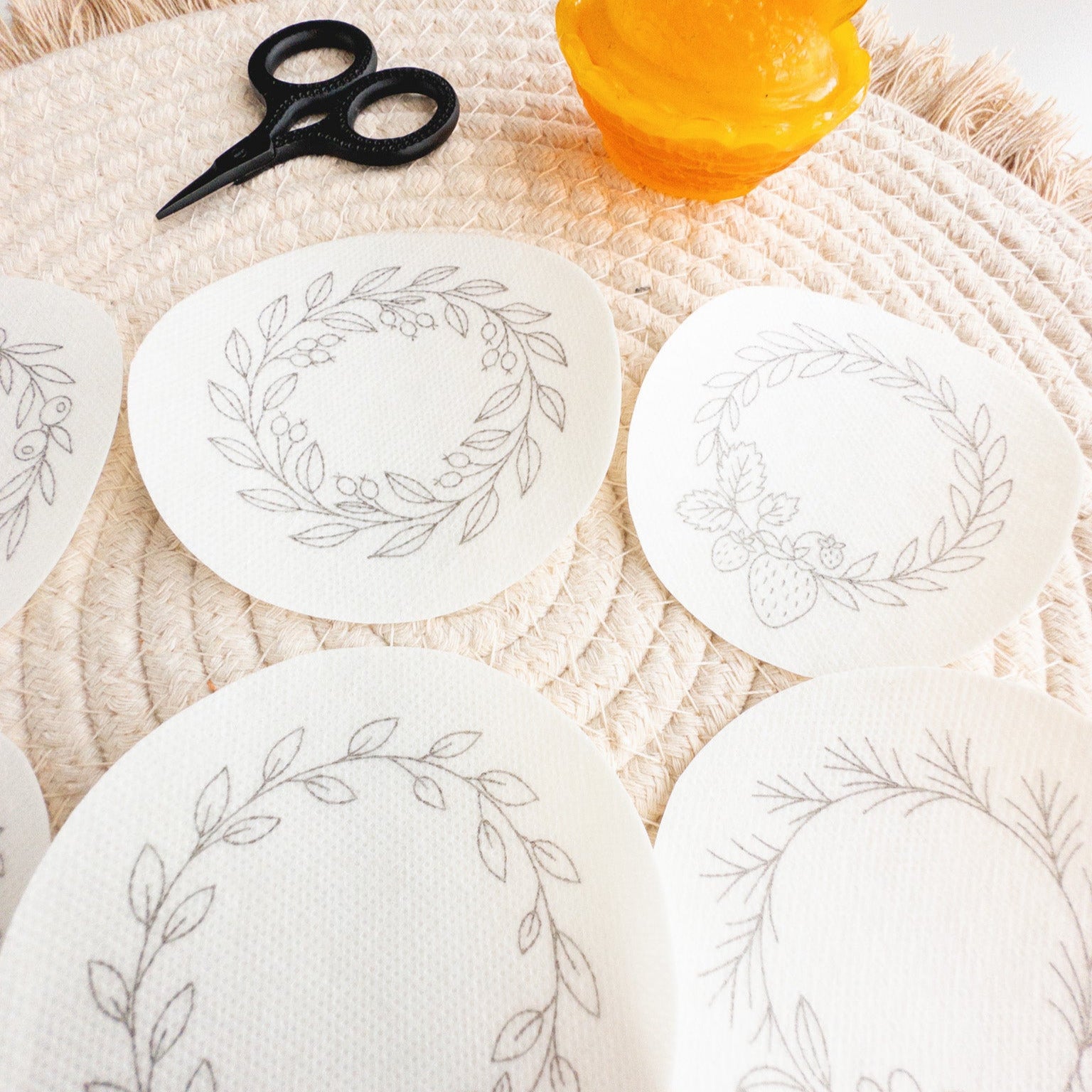 Six white oval wash-away embroidery stickers with delicate gray fruit wreath designs on a woven mat.
