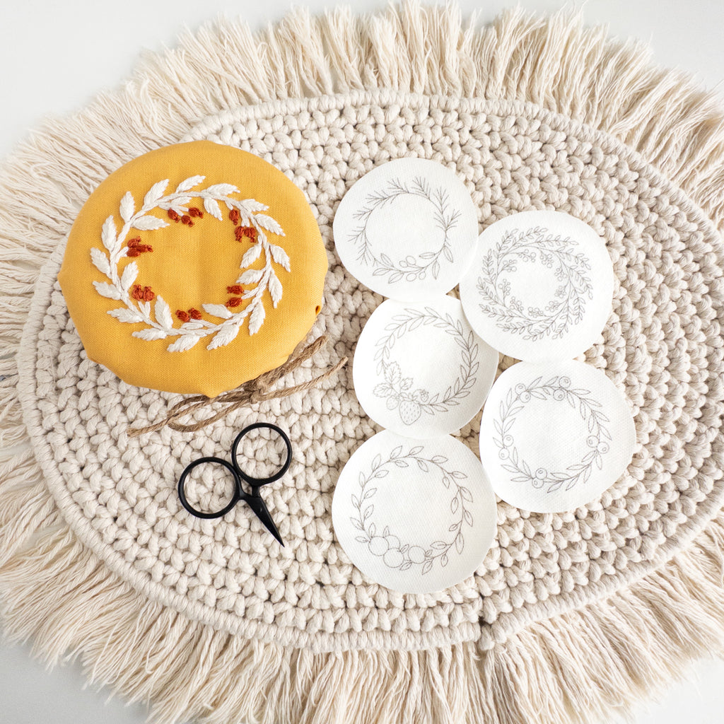 Embroidered fruit wreath on yellow fabric, with five wash-away patterns and small black scissors on a macrame mat.